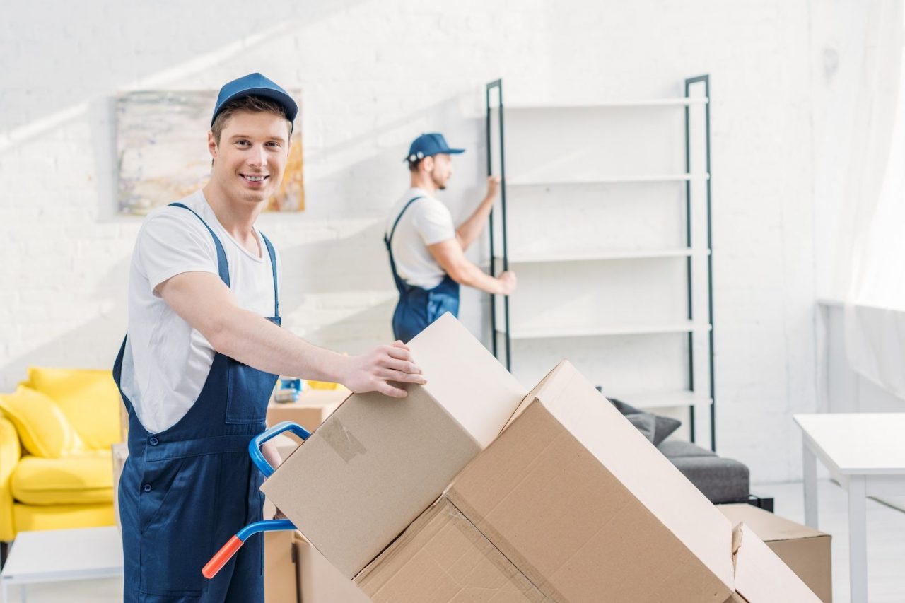two movers in uniform transporting cardboard boxes and furniture in apartment e1670221445352